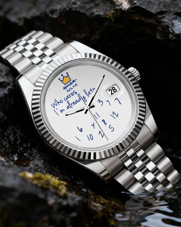 A silver stainless steel wristwatch with a white dial and blue hands, resting on dark volcanic rocks outdoors.
