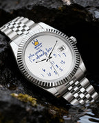 A silver stainless steel wristwatch with a white dial and blue hands, resting on dark volcanic rocks outdoors.